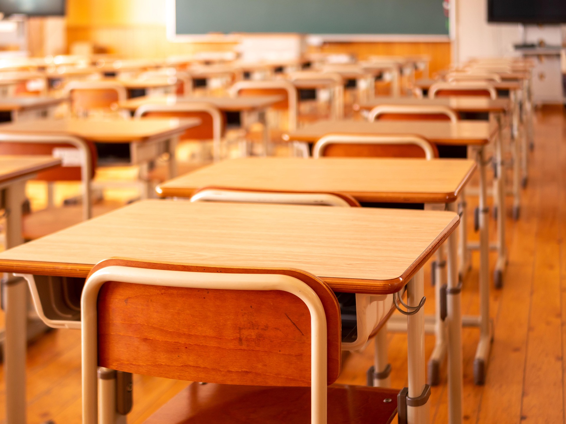 An empty classroom at the end of the day: rows of wooden desks and chairs receding towards a green chalkboard, washed in warm late-afternoon light.